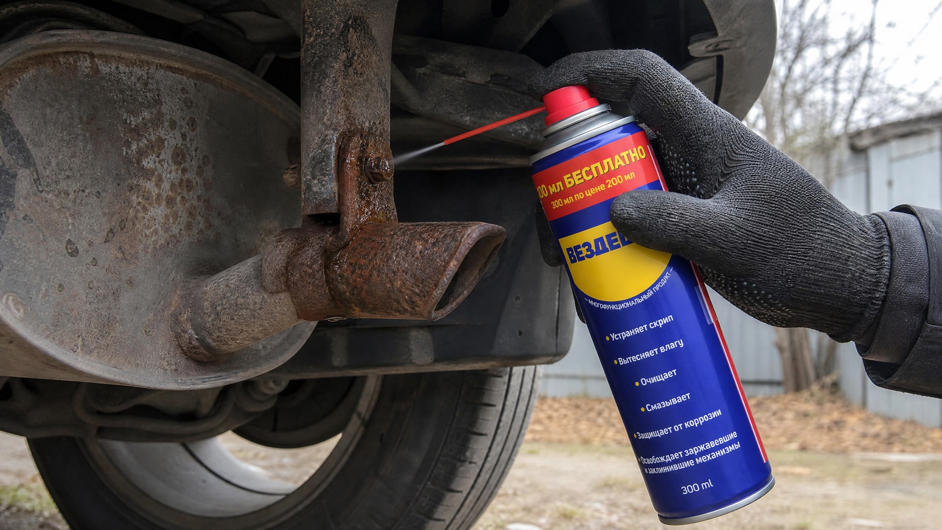 Рука в перчатке наносит универсальную смазку WD-40 на болты глушителя.