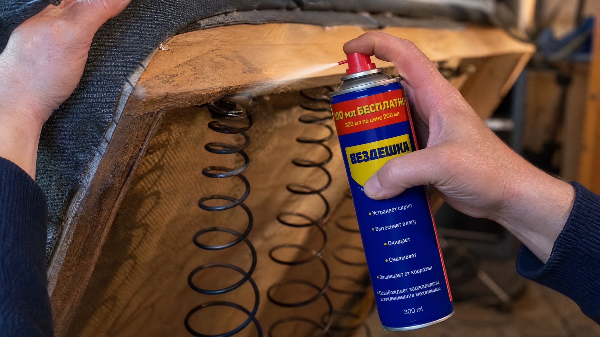 Рука в перчатке наносит WD-40 на скрипящую пружину дивана.